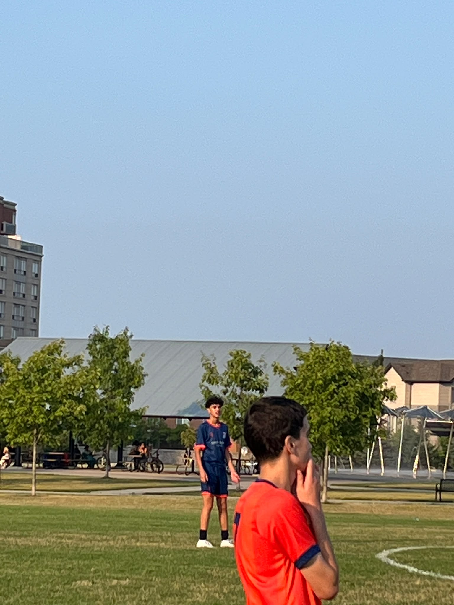Two young footballers pause on a grass field in a Toronto park, with trees and city buildings behind them