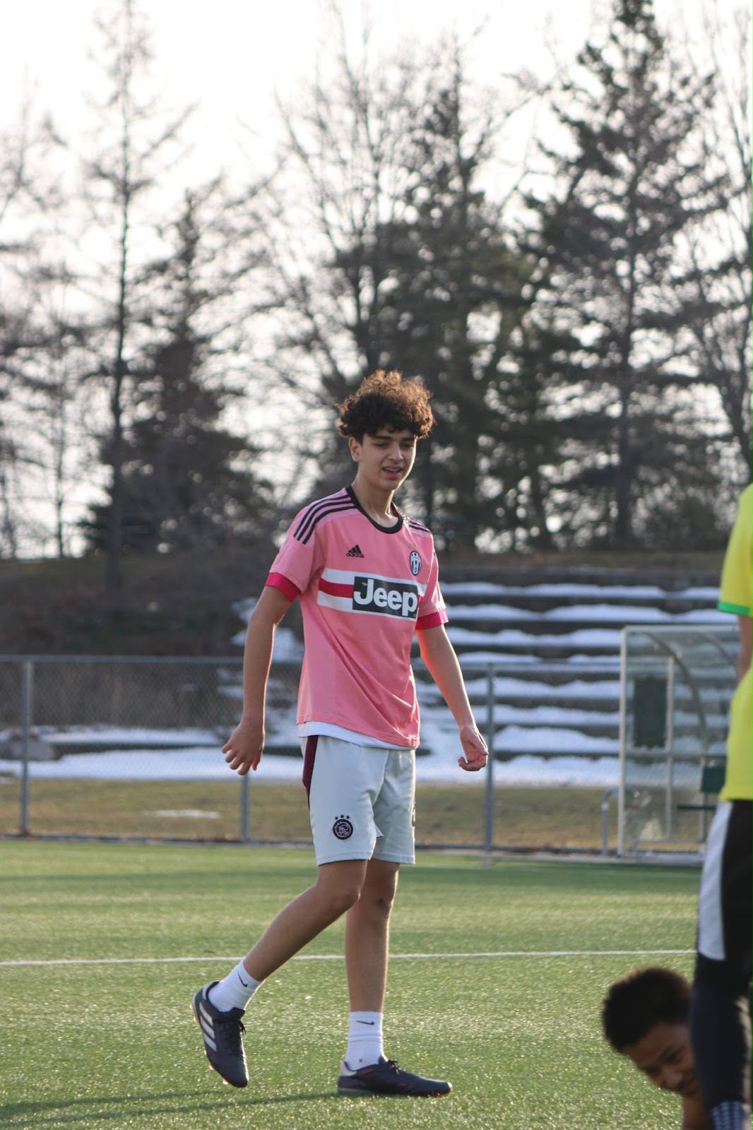 A young player in a pink training top walks across an artificial turf pitch in Toronto with snow-dusted bleachers behind
