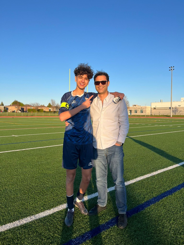A young football captain stands with a family member on an artificial turf pitch in Toronto, smiling together after training