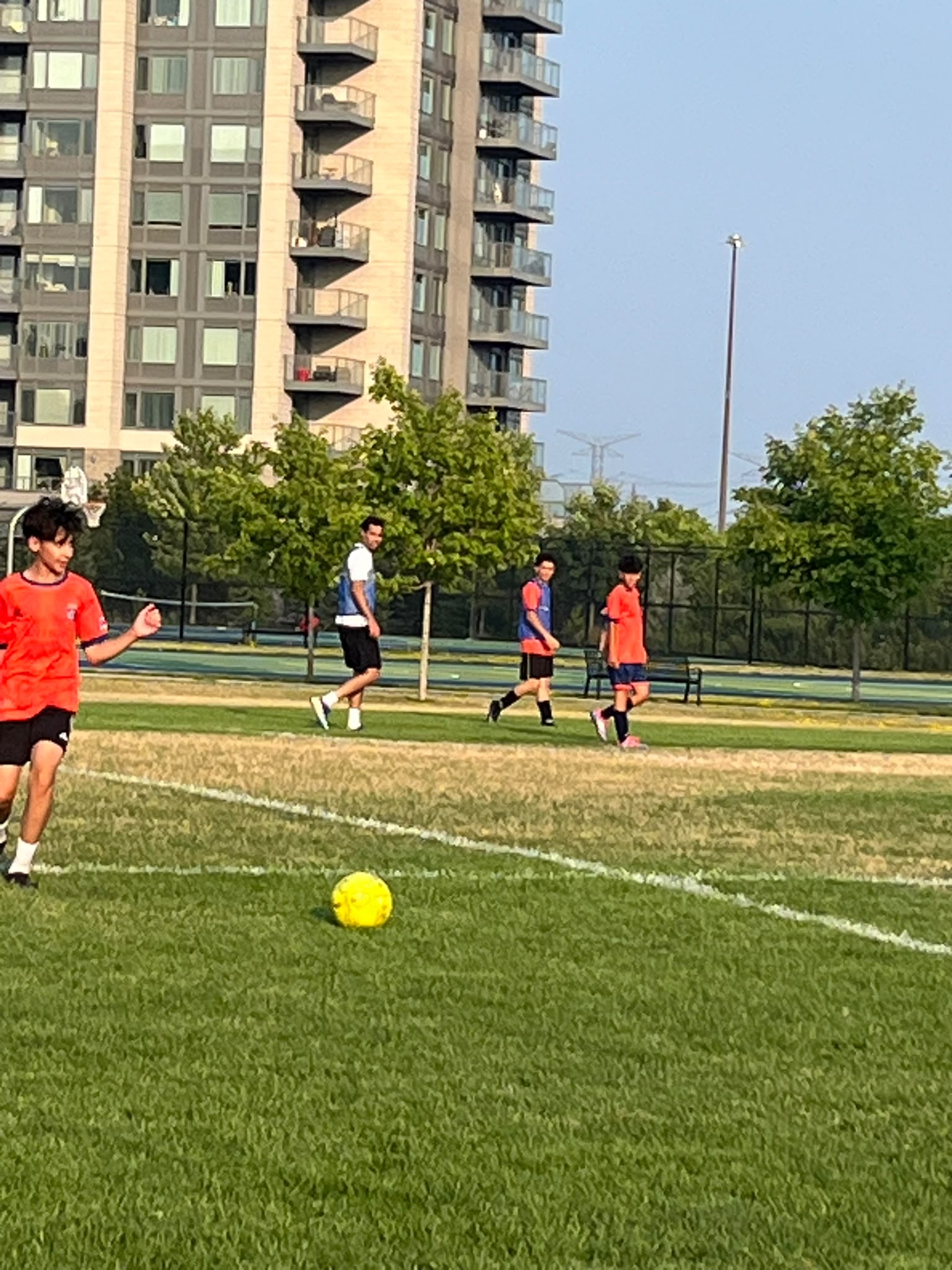 Young footballers train on a grass pitch in a Toronto park, with apartment towers rising behind the tree line